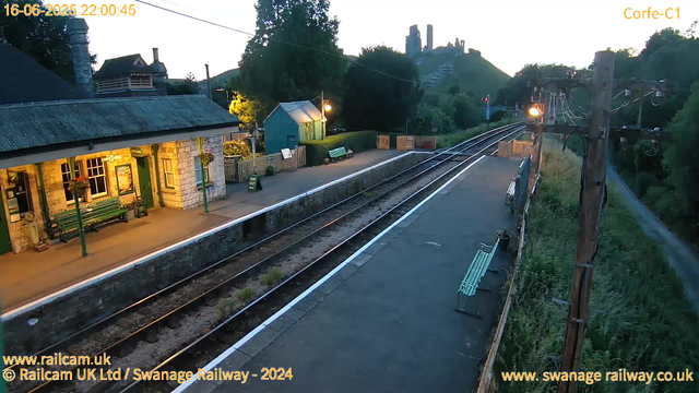 A small railway station platform at dusk, with a building on the left illuminated by warm yellow lights, benches, and potted plants. Two train tracks run parallel through the scene, and there is a footpath with a bench and some greenery on the right. In the background, hills are topped with ruins or castle remnants, and an electric pole with wires is visible on the right side. The timestamp on the image indicates it was taken at 22:00 on June 16, 2025.