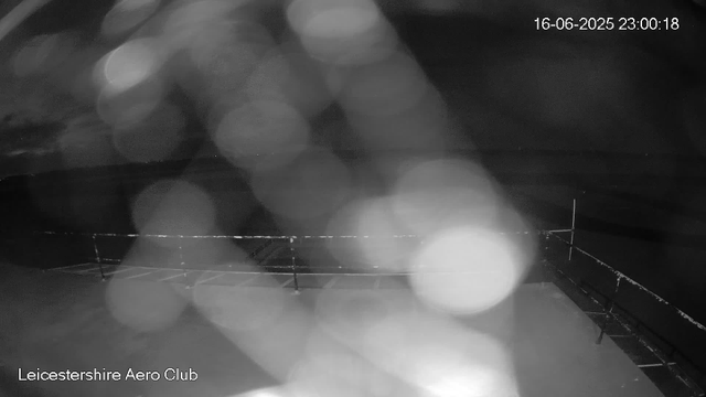 A black-and-white webcam image showing a cloudy, rainy night sky over a flat landscape. Raindrops are causing a blurred effect on the camera lens, and some illuminated bokeh lights are visible. The image has a timestamp of 16-06-2025 at 23:00:18 and is labeled "Leicestershire Aero Club" at the bottom left corner.