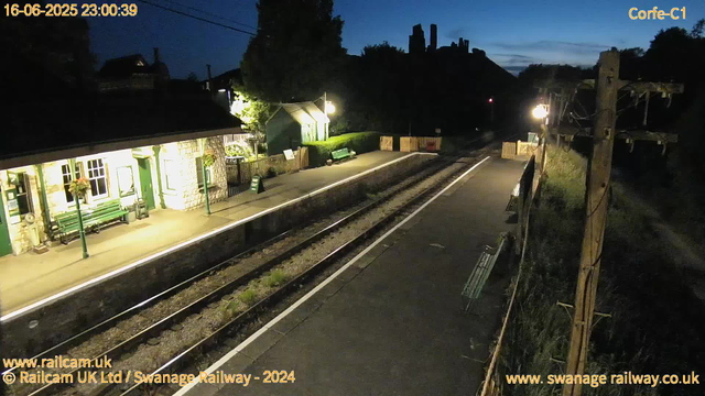 A small train station platform at dusk or early evening, illuminated by a few street lamps. The platform has a green bench, potted plants, and flower arrangements near the windows of a stone building. There are two train tracks running parallel, with some grass and weeds growing between the rails. In the background, there are trees and a darkening sky with faint blue hues. On the right side, there is a wooden utility pole with wires and a few benches attached. The image has timestamps and website credits overlaid.