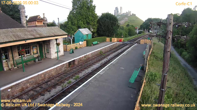 A small train station platform with two green benches, a stone building with a green door and hanging flower baskets, and a pathway on the right side separated by a wooden fence. There are railway tracks in the middle, with a green shed behind the platform. In the background, there are lush trees, a hill with a castle or fort ruins on top, and some buildings amidst the greenery. Power lines and a wooden utility pole are also visible on the right side. The scene appears to be early morning, as indicated by the timestamp.