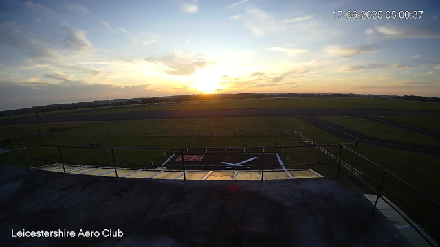 A sunrise over an open field at Leicestershire Aero Club, with the sky partly cloudy and the sun just above the horizon. In the foreground, there is a helipad with a large white "X" marked on it, and surrounding safety fencing. The landscape extends into grassy fields and a runway in the distance. The image includes a timestamp in the top right corner showing the date as June 17, 2025, and the time as 05:00:37.