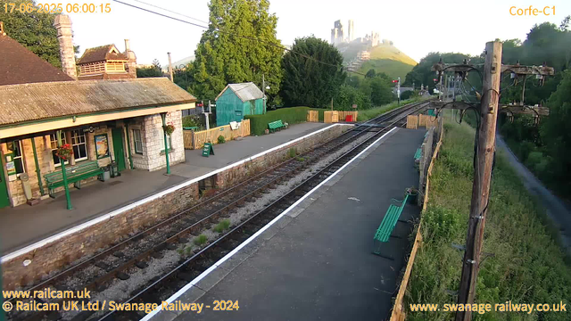 A small railway station platform with a building on the left that has green benches, hanging flower baskets, and a sign indicating "CORFE-CASTLE." There are flower baskets hanging from the building, and a sign saying "WAY OUT" near the platform edge. The railway tracks run through the center, with some plants growing between the sleepers. To the right, there are wooden fences, green benches, a small green shed, and a mature tree. In the background, there is a lush green hill with a castle visible atop, under a clear sky. An old wooden utility pole with wires stands near the platform edge on the right side. The timestamp indicates the morning of June 17, 2025.