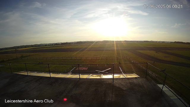 A wide view of Leicestershire Aero Club taken during sunrise, with the sun low on the horizon casting bright rays over an open grassy airfield. The sky is partly cloudy with soft sunlight illuminating the scene. In the foreground, there is a small helipad with a red "H" marking, surrounded by a metal railing. The landscape extends into flat, grassy fields and a runway in the distance. The timestamp in the top right corner shows the date and time as June 17, 2025, at 6:00 AM. The bottom left corner displays the text "Leicestershire Aero Club."