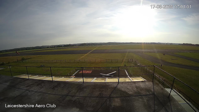 An outdoor view of an airfield on a sunny day, with a bright sun in the sky and some scattered clouds. There is a large grassy area with a paved runway stretching across the background. In the foreground, part of a building or observation deck is visible with a railing and what appears to be a windsock on a pole. The image has a timestamp showing it was taken on June 17, 2025, at 7:01 AM. The text "Leicestershire Aero Club" is displayed at the bottom left corner.