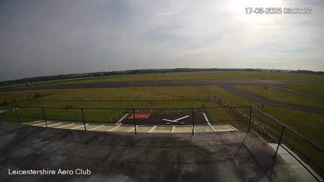 An empty airfield with a large clock on the ground displaying the time as approximately 4:47. A windsock is visible on the left side, and a fence surrounds the clock. In the background, there are runways and grassy areas extending to the horizon under a partly cloudy sky. The timestamp in the upper right corner reads "17-06-2025 08:00:26," and the bottom left corner indicates the location as "Leicestershire Aero Club."