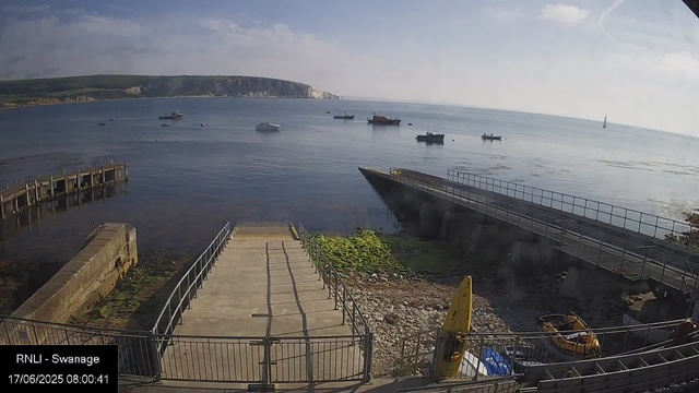 A calm seaside scene with a pier extending into the water and various boats anchored nearby. In the foreground, there is a paved walkway and a ramp leading down to the rocky shoreline, with some equipment and a yellow buoy visible. In the background, there are cliffs with a partly cloudy sky overhead.