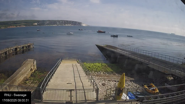 A scenic view of a harbor with calm water and several boats floating. In the foreground, there is a concrete pier with railings, some green algae on the rocks, and a small yellow warning sign. To the right, a section of the shoreline has yellow and black flotation devices. In the distance, white cliffs and green land are visible under a partly cloudy sky. The timestamp at the bottom left reads: "17/06/2025 09:00:31."