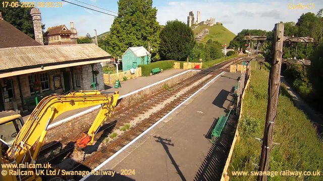 A small train station platform with a railway track running through the center. On the left, there is a stone building with a tiled roof and a chimney. A yellow excavator is parked along the platform, partially visible in the lower left corner. A green bench and a turquoise shed are on the far side of the platform. Several green benches are along the edge of the platform, with some flowers hanging below a window. To the right, a tall wooden pole with wires extends from the foreground to the background. In the distance, a hill with what looks like castle ruins or old stone structures sitting atop it, surrounded by trees and greenery. The sky is clear with a few clouds.
