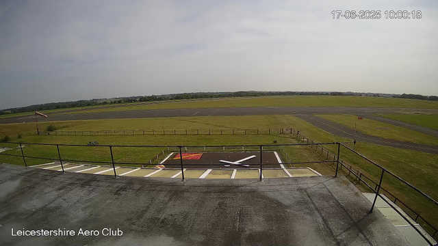 A widescreen view of an empty runway and taxiway at Leicestershire Aero Club. The image is taken from a high vantage point on a building's balcony or observation area. In the foreground, there is a helicopter landing pad with a large white "X" marking it and a red and white sign nearby. To the left, there is a flagpole with a windsock. The sky is partly cloudy with some sunlight, and the time and date are displayed in the upper right corner as "17-06-2025 10:00:18." The bottom left corner contains the text "Leicestershire Aero Club."
