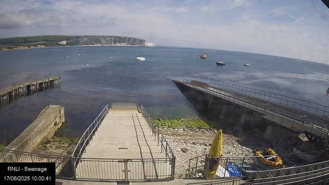 A tranquil seaside scene showing a small pier extending into calm water, with boats anchored nearby. In the background, white cliffs rise against a partly cloudy sky. A concrete ramp leads down to the water, and there is some rocky and algae-covered shore below the ramp. A yellow rescue boat and other equipment are stored near the shoreline, with a metal railing surrounding the area. The scene appears to be during the day with bright, natural light.
