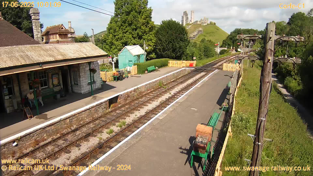 A small train station platform with a building on the left side, featuring a chimney, a bench with two people sitting, and a green bench nearby. There are two sets of railway tracks running through the center of the image, with some plants growing between the tracks. On the right side, there are green benches, a wooden fence, and a row of utility poles with wires. In the background, a green hill with trees and a castle ruins atop it can be seen under a partly cloudy sky. There is some construction fencing in yellow and red near the tracks. The timestamp in the top left corner indicates the date and time as June 17, 2025, at 10:01:09.