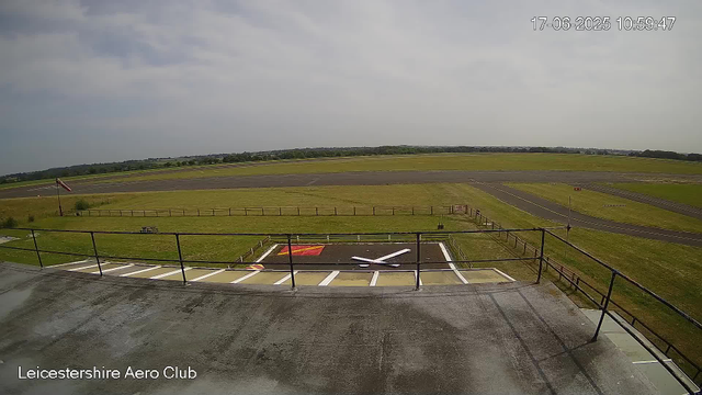 A view from a webcam at Leicestershire Aero Club showing a grassy airfield with a small helipad in the foreground. The helipad has a white "H" in the center and is surrounded by a black railing, with a red and yellow marking nearby. The background features an expansive flat landscape with runways, green fields, and a cloudy sky. The timestamp in the top right corner shows the date and time as 17-06-2025, 10:59:47.