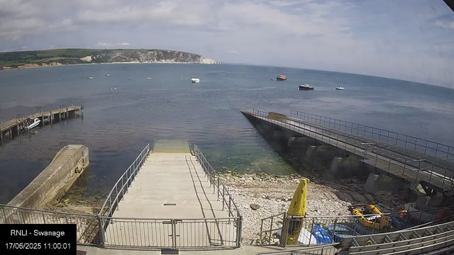 A view of a marina with a concrete boat ramp leading into the water. Several small boats are docked along the piers on the left, and a few boats are seen sailing in the distance across the calm sea. In the background, there are white chalk cliffs and green hills under a partly cloudy sky. There are safety signs and equipment, including yellow caution cones and life vests, near the shoreline. The timestamp on the image indicates it was taken on June 17, 2025, at 11:00:01.