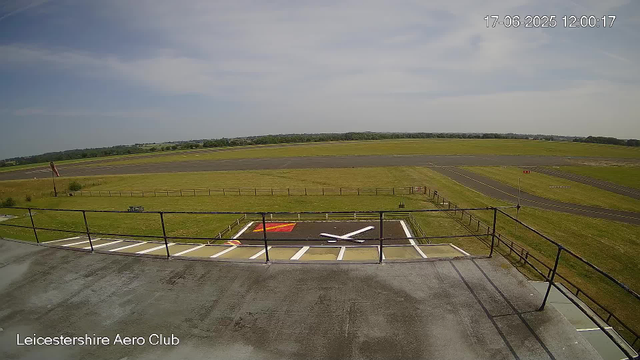 An empty observation deck at Leicesterhire Aero Club overlooks an aprons and taxiway area of an airfield with grassy fields and trees in the distance. The sky is partly cloudy with patches of blue. The current date and time is visible in the upper right corner: 17-06-2025 12:00:17.
