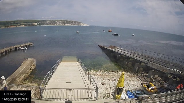 A view of a harbor with several small boats floating on calm water, a pier extending into the water on the left, and a newly constructed or repaired dock on the right. In the foreground, a person wearing a hat and light clothing stands near the fence, and some safety equipment, including a yellow life raft and a yellow warning sign, are visible. The sky is partly cloudy with a mix of clouds and blue sky, and a distant coastline with cliffs and green hills can be seen across the water.
