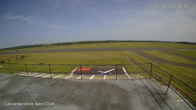An empty aircraft parking area with a helicopter landing pad marked with a large white "X" on the ground. The pad is bordered by a black metal railing, and there are white and red markings. Beyond the pad, there's a grassy field with a small windsock on the left side, and a long runway in the distance under a clear blue sky with a few scattered clouds. The timestamp in the top right corner reads "17-06-2025 13:00:15," and the bottom left corner says "Leicestershire Aero Club."