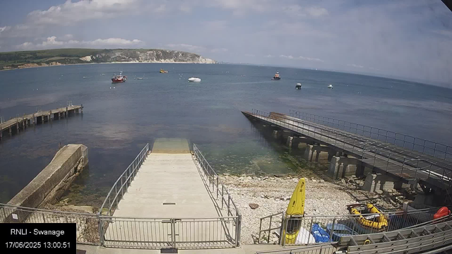 A view of a seaside dock area with calm water and several boats anchored offshore. The sky is partly cloudy, and in the background, there are white chalk cliffs and green land. In the foreground, there are two jetties, one with a ramp leading into the water, and the other a platform with a railing. There are also some yellow and orange safety equipment and a yellow inflatable boat on the rocky shore. The timestamp indicates the image was captured on June 17, 2025, at 1:00 pm.