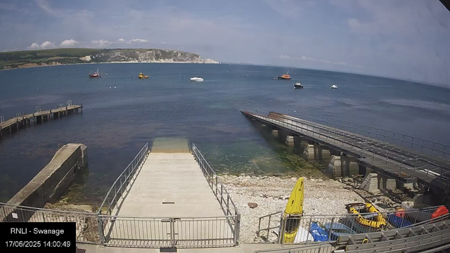 A view of a harbor with a concrete and wooden dock extending into the water, surrounded by metal railings. Several boats are anchored in the distance, and a grassy hill with white cliffs is visible on the horizon under a partly cloudy sky. There are some yellow and red rescue boats and equipment near the shoreline. The timestamp reads "17/06/2025 14:00:49" and the location is labeled as RNLI - Swanage.