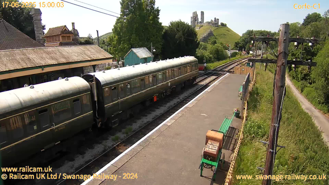 A vintage train with dark green carriages is stopped at a small rural station platform. The platform has a green bench, a potted plant, and a small luggage trolley. To the right, there are wooden utility poles with wires stretching overhead. In the background, a lush green hill with the ruins of an ancient castle on top can be seen, along with trees, houses, and a clear blue sky.