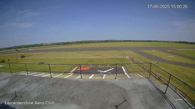 An outdoor view from a webcam at Leicestershire Aero Club showing a wide, clear sky with a few small clouds, a runway, and surrounding grassy fields. A small aircraft is taxiing on the runway in the distance. The image is timestamped June 17, 2025, at 3:00 PM.
