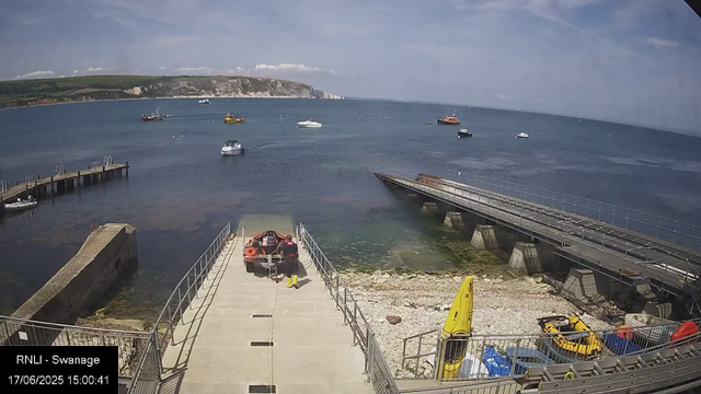 A view of a harbor with several boats floating on calm water, a rocky shoreline, and a pier extending into the water. In the foreground, there is a small dock with a person standing next to a boat, wearing a life jacket and yellow boots. Nearby, there is a yellow rescue board and a small inflatable boat. The background shows a green hillside and partly cloudy sky. The image has a timestamp of 17/06/2025 at 15:00:41 and is labeled "RNLI - Swanage."