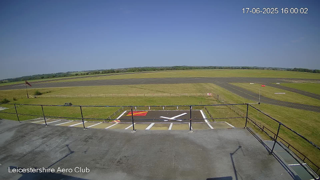 An empty aircraft parking area with a helipad marked by a large white "H" and a red square. The viewpoint is from a camera mounted on a platform or roof, overlooking the tarmac and surrounding grassy fields. The background features open skies with a clear blue horizon, and a windsock is visible on the left side. The timestamp shows the date as June 17, 2025, at 4:00 PM. The location is identified as Leicestershire Aero Club.