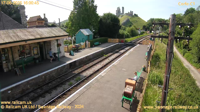 A small train station platform is visible with a stone building on the left, featuring a porch and a person standing near a bicycle. There are green benches along the platform, and a woman in a white top and hat is sitting on one of them. To the right, there are wooden utility poles with wires, and a narrow path runs parallel to the railway tracks, which extend into the distance towards a hill with ruins or castle remnants at the top. There are trees and greenery surrounding the area, with a small turquoise shed near the station. The sky is clear and sunny.