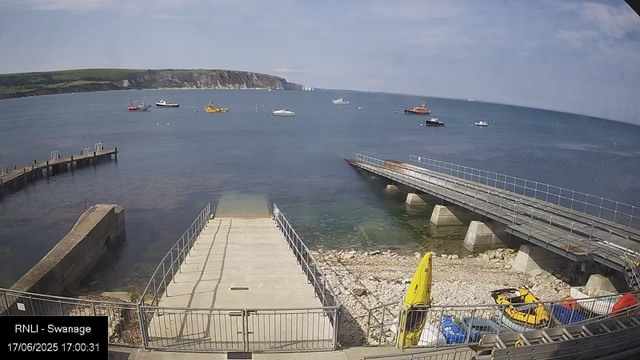 A view of a harbor with several boats anchored in the water. In the foreground, there is a concrete pier with metal railings, a small set of stairs leading down into the water, and a concrete path. To the right, a ramp extends over the water, supported by concrete pillars. On the rocky shore area below, there are yellow kayaks, a blue and red pedal boat, and other boating equipment. The background features a hilly coastline with white cliffs and a partly cloudy sky.