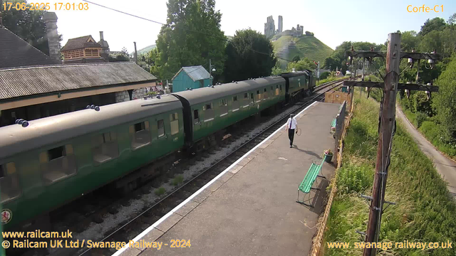 A green passenger train is stopped at a small rural station platform. A man dressed in a white shirt and dark trousers is walking along the platform carrying a hat or cap in one hand and a bag in the other. There are green benches and potted plants along the platform, with a wooden fence separating the platform from a grassy area. On the right, there are old wooden utility poles with wires. In the background, there are trees, small houses with tiled roofs, and an ancient stone castle on a hill. The weather is sunny and clear. The image has a timestamp of June 17, 2025, at 17:01.