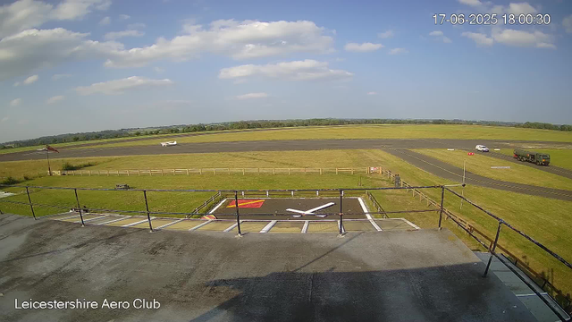 A view of a sunny day at Leicesterhire Aero Club showing an airfield with a runway and surrounding grassy areas. There is a white aircraft parked on the grass and several vehicles driving on the taxiway. A windsock is visible on the left side, and a viewing platform with a railing is in the foreground. The timestamp in the top right corner indicates the date and time as 17-06-2025 18:00:30.