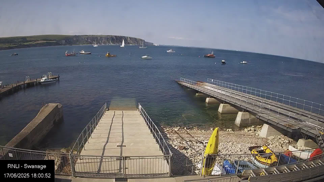 A view of a harbor with a concrete pier extending into calm water, boats floating on the surface, and a hilly coastline in the background under a partly cloudy sky. There are yellow and blue rescue or lifeguard equipment and a few small vehicles at the front.