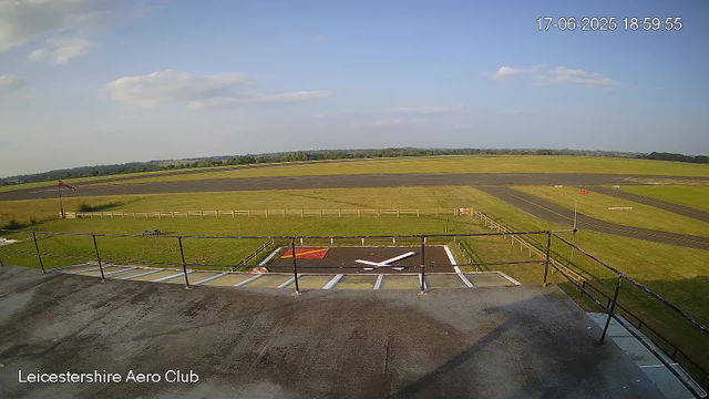 An empty airfield with a small helipad area in the foreground, surrounded by a fence. The background shows a wide expanse of open grass and paved taxiways or runways under a partly cloudy sky. The timestamp indicates the date and time as June 17, 2025, at 18:59:55. The bottom left corner displays the text "Leicestershire Aero Club."