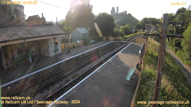 A small railway station platform with a stone building on the left, a person standing near the entrance. The railway tracks run straight ahead, with electric poles on the right side. There are green benches along the platform, and the sun is shining, creating lens flares in the image. In the background, there are hills with trees and a castle at the top, along with some houses and greenery. The image is bathed in warm sunlight, suggesting it is late in the day.