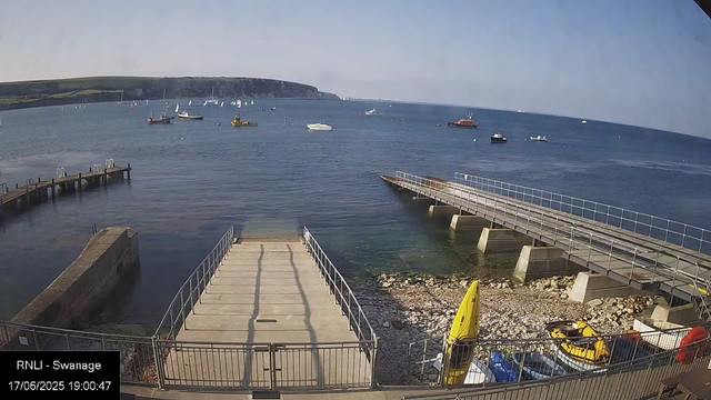 A view of a harbor with a dock extending into calm water, boats floating in the distance, and a rocky shoreline. There are two walkways with railings leading into the water, and some yellow boats and equipment are visible on the rocky shore in the foreground. A headland with cliffs and vegetation can be seen in the background under a clear sky.