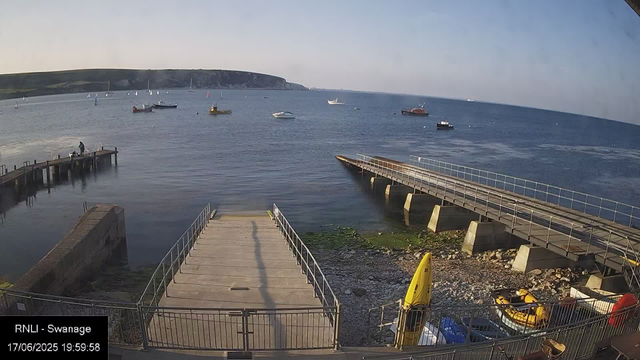 A view of a harbor with several boats on calm water. There is a wooden walkway leading into the water on the left and a concrete pier extending out on the right. In the foreground, there are yellow rescue boats and some equipment. A person is standing on the wooden pier, and the background features a hilly coastline under a clear sky. The timestamp indicates the date as June 17, 2025, at 19:59:58, and the location is labeled as RNLI - Swanage.