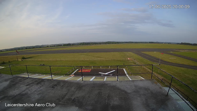 An overcast sky over a grassy airfield with taxiways and runways stretching into the distance. In the foreground, there is a helipad marked with a large white helicopter symbol and a red square, surrounded by a railing. A windsock is visible on a pole near the left side, and a small wooden bench is positioned nearby. The image is labeled "Leicestershire Aero Club" in the bottom left corner, with the date and time stamped in the top right corner as "17-06-2025 20:00:09".