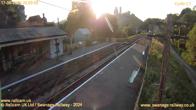 A railway station platform at sunset with empty benches, a small building, and overhead electric lines. The tracks extend into the distance, and there is a hill with castle ruins in the background. The sun is low in the sky, casting warm light and lens flare effects.