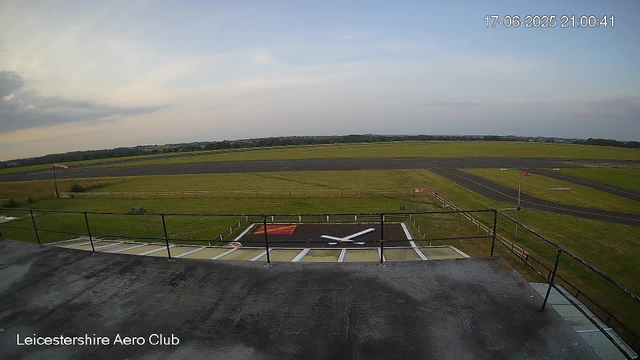 A view of an airfield with a runway and grassy areas, taken from a webcam at Leicesterhire Aero Club. In the foreground, there is a helipad with a white helicopter symbol and a red section. The sky is partly cloudy, and the time and date displayed are 17-06-2025 at 21:00:41.