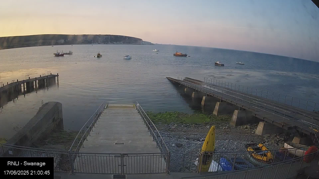 A scenic view of a coastal area during sunset or sunrise. In the foreground, there are two concrete piers extending into the water with metal railings, a yellow kayak, and some parked cars. The middle ground features a calm body of water with a few boats scattered across the surface. The background presents a distant shoreline with steep cliffs under a pastel sky. The image appears to be captured from a webcam.