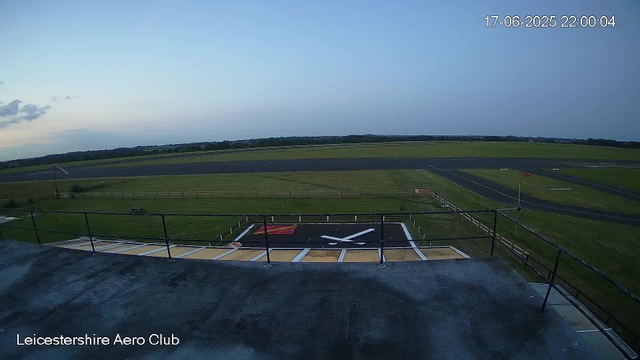 An outdoor view from a webcam at Leicestershire Aero Club shows a clear sky at dusk. In the foreground, there is a helipad painted black with a white circle and a red square, and a large white clock symbol. Surrounding the helipad is a dark paved area and a metal railing. Further back, a grassy field extends to a paved runway, with a few small white structures and red and white markings along the runway. A British flag is flying on a flagpole to the left, and a parking or picnic area with a bench is visible nearby. The timestamp in the top right corner indicates the time and date as 17-06-2025 at 22:00:04. The bottom left corner displays the text "Leicestershire Aero Club."