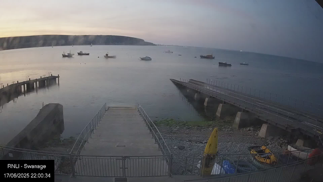 A view of a harbor at dusk with a calm sea and several boats anchored in the water. In the foreground, there is a concrete pier with metal railings and steps leading into the water, along with a yellow caution sign and some parked yellow and gray vehicles. A larger platform extends out into the water on the right. The background features cliffs along the horizon under a partly cloudy sky.