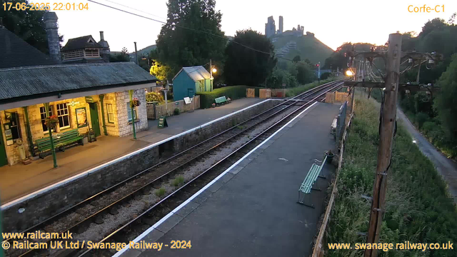 A quiet railway station platform at dusk, with a small stone building on the left side illuminated by warm light, featuring green benches and flower baskets hanging from lamp posts. There are train tracks extending into the distance, surrounded by greenery and hills, with a large hilltop castle or ruins visible in the background. A wooden utility pole with wires runs along the right side of the platform, which is bordered by a fence and a narrow pathway. The sky has a fading light, indicating evening time.