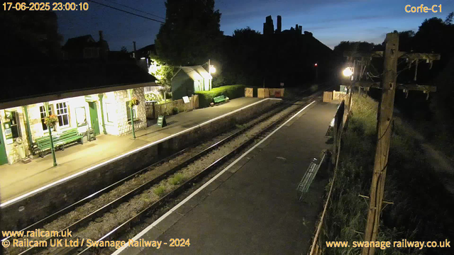 The image shows a small railway station platform at dusk or early night. The station building on the left is illuminated with warm lighting and has hanging flower baskets outside the windows. A green bench and a small shed are visible further along the platform. The railway tracks run through the center of the image, with a wooden fence and some fencing barriers at the far end. On the right side, there are utility poles with wires, and the sky is darkening, with a hint of blue. The timestamp on the top left indicates the date and time as 17-06-2025, 23:00:10.