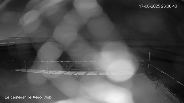A black and white night-time webcam image shows an empty airstrip or runway with a safety barrier at the edge. The sky appears dark, suggesting late night or early morning. The image has a timestamp in the upper right corner reading "17-06-2025 23:00:40," and the words "Leicestershire Aero Club" are displayed at the bottom left corner. The scene is blurry with visible bokeh, possibly due to condensation, rain, or camera focus issues.