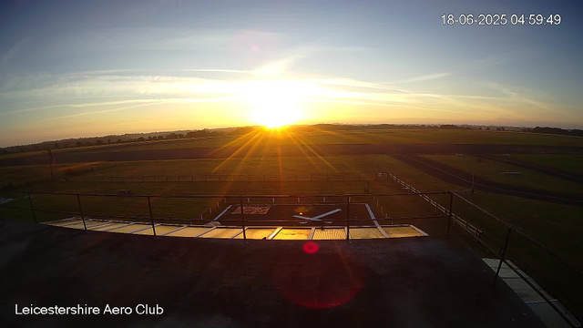 A sunset view over an open field with a clear sky, showing the sun near the horizon casting a golden light. In the foreground, there is a small fenced area with a windsock and some equipment. The scene is labeled "Leicestershire Aero Club" and the timestamp indicates it was taken on June 18, 2025, at 4:59 AM.