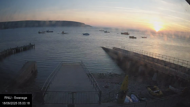 A scenic view of a harbor at sunrise, with the sun just above the horizon casting a golden glow over the water. Several boats are anchored in the distance, and a wooden pier extends into the water on the left side of the image. In the foreground, a small boat is on a platform, and a yellow caution sign is visible near the bottom right. The sky is partly cloudy, with streaks of clouds and faint contrails. The landscape features a distant headland or cliff along the horizon.