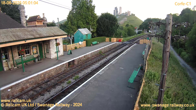 A small railway station platform with a stone building, green benches, and flower baskets hanging from lampposts. The platform is empty, with a "Way Out" sign near the waiting area. Rolling green hills and a castle ruin are visible on a hill in the background, with some trees and a small shed next to the platform. A road runs parallel to the railway tracks, separated by a grassy verge and wooden fencing. An old wooden pole with electrical wires crosses the scene on the right side. The timestamp shows the date and time as 18-06-2025 05:01:04.
