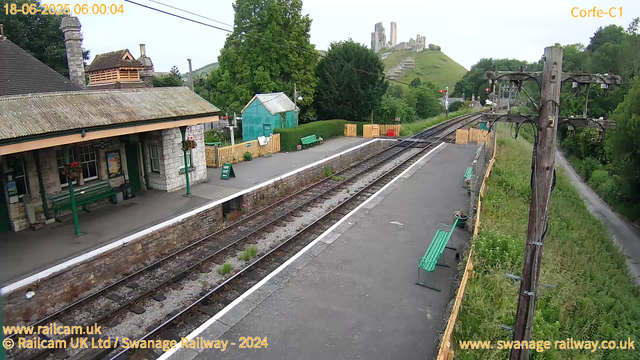 A small railway station platform with a building on the left side, featuring a pitched roof, stone walls, and hanging flower baskets. There are several green benches, some flower pots, and a "Way Out" sign. The railway tracks run parallel to the platform, with a countryside landscape of trees and grass beyond. In the background, on a hill, there are castle ruins. On the right side, there are tall wooden utility poles with electrical wires, and a narrow green pathway runs alongside the structure. The timestamp shows the date and time as 18-06-2025 06:00:04.