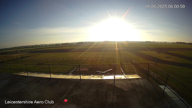 The image shows a bright, sunny morning at Leicestershire Aero Club with the sun shining intensely in the sky, creating a glare and lens flare effects. The scene includes a grassy field with a few small structures in the foreground, surrounded by a safety fence. The background features open, flat terrain with patches of grass and a distant tree line on the horizon. The timestamp in the top right corner indicates the date and time as June 18, 2025, at 6:00 AM.