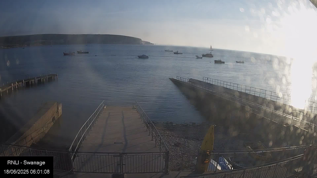 A view of a calm harbor with several boats anchored in the water. There is a pier extending into the sea on the left and a ramp leading into the water on the right. The sky is clear with the sun shining brightly, creating reflections on the surface of the water. In the background, there are distant hills or cliffs along the shoreline. A yellow kayak or small boat is visible near the bottom right corner. The timestamp indicates the image was taken early in the morning.